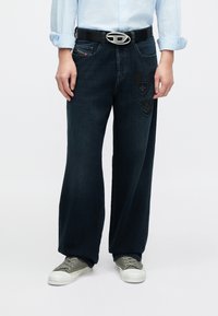 Sötét farmer, bőszárú nadrágok díszítő foltokkal és varrással az elején. Fekete övvel és zöld tornacipővel viselve. Világoskék ing.