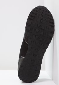 Czarna podeszwa sportowego buta z teksturowanej gumy i trójkątnym wzorem bieżnika, z szarym, plecionym wierzchem i gładkim obcasem.