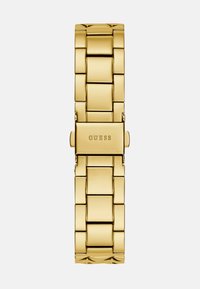 Kultainen metallikello, jossa on suorakulmainen kasvopinta, linkitettu ranneke ja solki, jossa on brändinimi "GUESS". Tasainen pinta ja kiillotettu viimeistely.