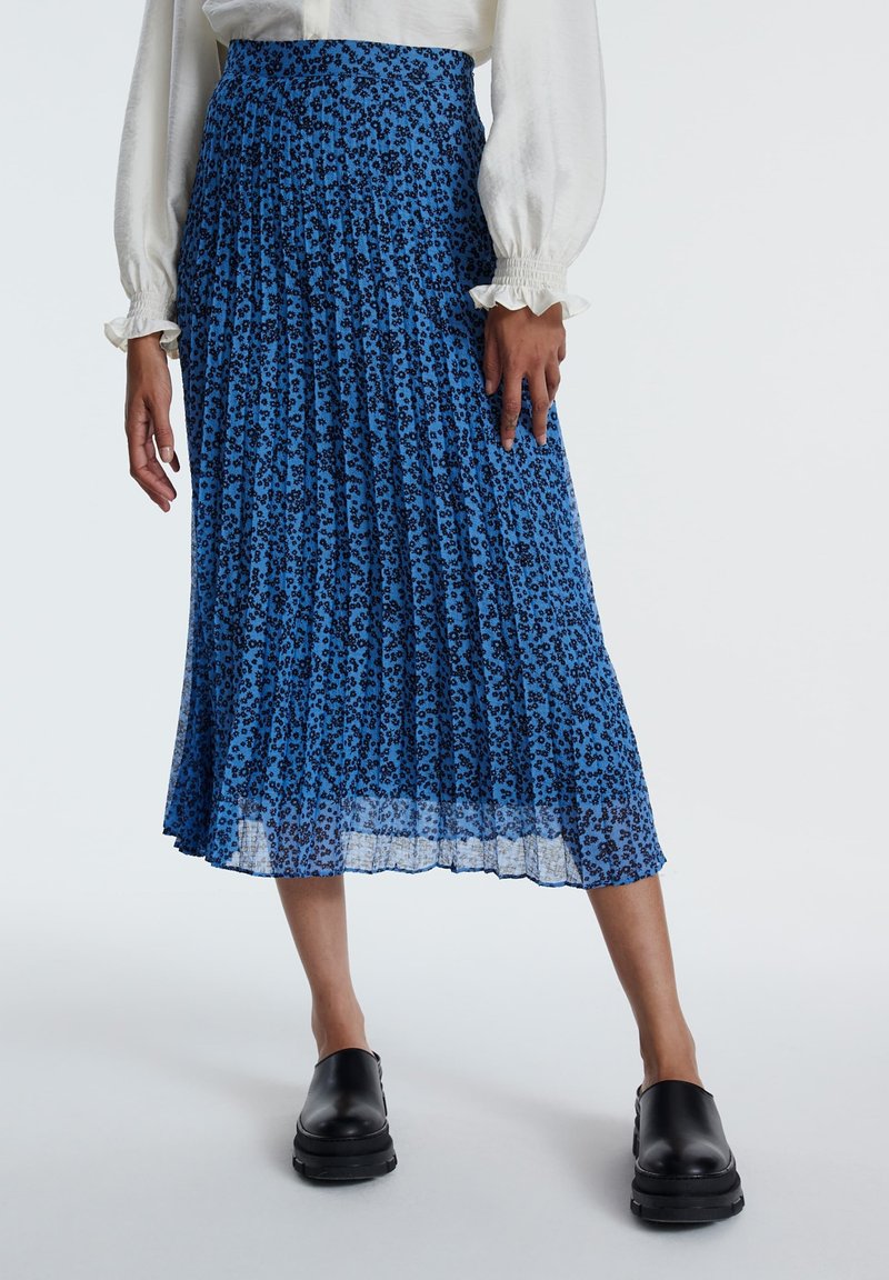 Blaue plissierte Midirock mit schwarzem Blumenmuster, transparenter Textur und einem gerafften Taillenbund, kombiniert mit schwarzen Slippern.
