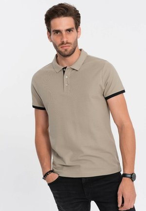 Camisa polo beige con una superficie texturizada, cuello negro y acentos en las mangas. Presenta una botonera de tres botones y mangas cortas. Llevada con pantalones negros.