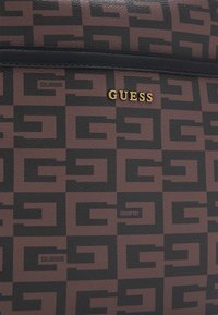 Materiale sintetico marrone con un motivo geometrico nero. Un emblema dorato "GUESS" è ben in evidenza nella parte superiore.