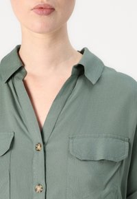 Camicia verde oliva con colletto, due tasche sul petto e una texture di tessuto morbida. Presenta un orlo arrotondato e bottoni trasparenti.