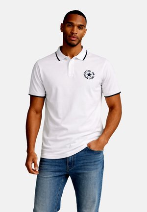 Man draagt een wit poloshirt met zwarte bies en een rond logo, gecombineerd met een blauwe spijkerbroek, staat met één hand in de zak.