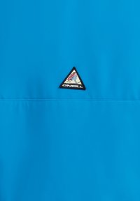 Zila ārējā apģērba daļa ar gludu auduma tekstūru, trīsstūrveida melnās un baltās krāsas logo uzlīme krūtīs un plakanām šuvēm.