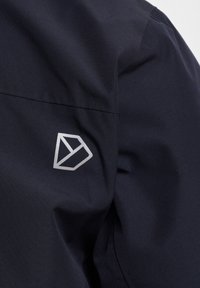 Tamsiai mėlynas švarkas su glotnia tekstūra, geometriniu sidabro logotipu ant rankovės ir struktūruotais siūlais, suteikiančiais puikiai pritaikytą išvaizdą.