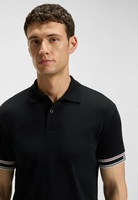 Svart polo t-shirt med knäppbar krage och ribbade ärmslut som har horisontella ränder i röd, vit och grön. Stryktålig tygtextur.