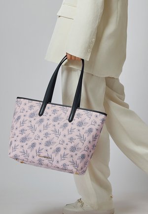 Persona con traje crema sosteniendo una gran bolsa tote rosa con un patrón floral negro y asas negras, caminando frente a un fondo plano.