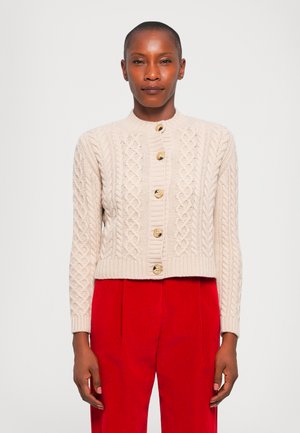 Gilet en tricot beige avec un motif en câble, col rond et cinq boutons en bois. Associé à un pantalon en velours côtelé rouge, style cropped.