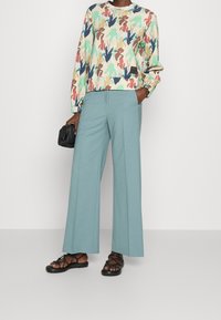 Sweat-shirt coloré à motifs avec manches longues, associé à un pantalon large bleu clair et des sandales noires à lanières, tenant un sac noir texturé.