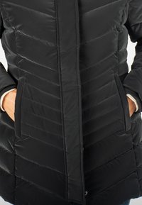 Manteau matelassé noir avec un motif en chevrons, doté de deux poches avant et d'une fermeture éclair. Fabriqué en matériau lisse et isolant.
