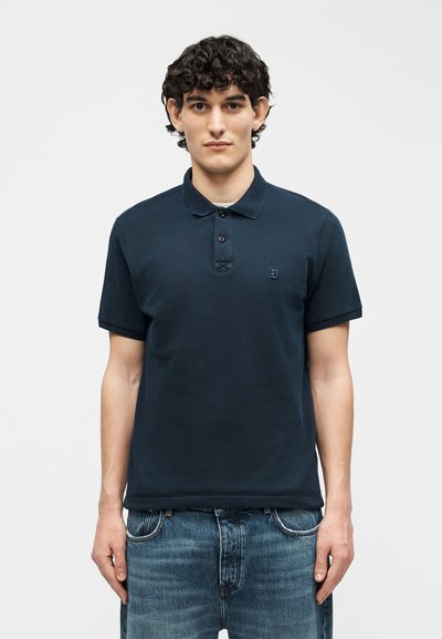 Jeune homme aux cheveux bouclés portant un polo bleu marine à manches courtes et un jean bleu, debout devant un fond clair uni.