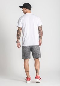 Bijela kratka majica s kratkim rukavima, sivi traper šortsevi s uzorkom, crna kapa i crvene tenisice s bijelim točkicama; vidljive tetovaže na rukama.