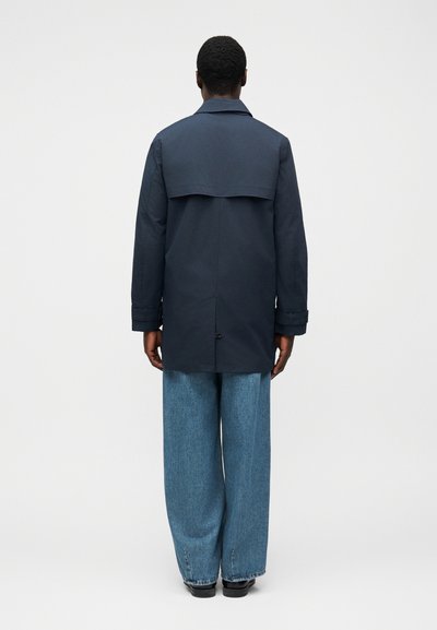 Σκούρο μπλε παρκά με οπίσθιο κάλυμμα, συνδυασμένο με ανοιχτό μπλε τζιν παντελόνι με φαρδιά μπατζάκια. Το ύφασμα φαίνεται λείο, με καθαρές γραμμές και ελάχιστες λεπτομέρειες.