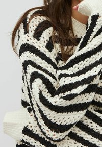 Pleťovaný svetr s chevronovým vzorem v černé a krémové barvě, s žebrovanými manžetami a otevřeným texturovaným designem.