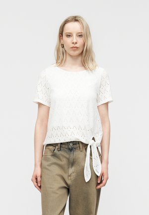 Vaaleahiuksinen nainen, jolla on valkoinen, silmukkakuvioinen lyhythihainen pusero solmittuna vyötärölle ja beiget korkeavyötäröiset farkut yksiväristä taustaa vasten.