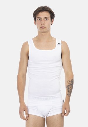 Jonge man met tatoeage op linker onderarm, draagt een wit tanktopje en witte onderbroek, staand tegen een effen lichte achtergrond.