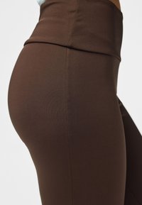Gros plan d'une personne portant des leggings marron taille haute, montrant la texture du tissu et la couture le long du côté et de la ceinture.