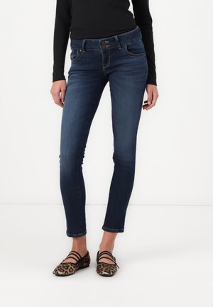 Woman wearing dark blue skinny jeans, black long-sleeve top, and leopard print flats with multiple straps, standing against a white background.