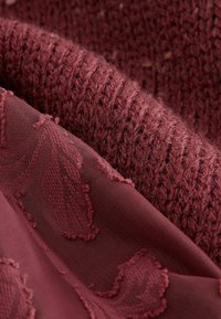Tissu bourgogne présentant une surface tricotée texturée et un revêtement lisse avec des motifs de feuilles. Douces variations dans le tissage et la profondeur des couleurs.