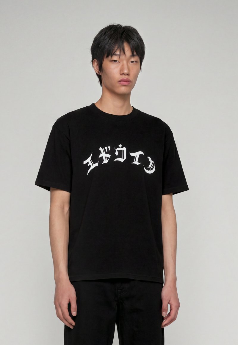 Jeune homme aux cheveux courts noirs portant un T-shirt noir avec un texte blanc stylisé sur la poitrine, debout devant un fond gris clair uni.