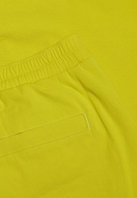 Zbliżenie na materiał w jasnej zieleni limonki, pokazujący elastyczny pas i prostokątną kieszeń z widocznym przeszyciem.