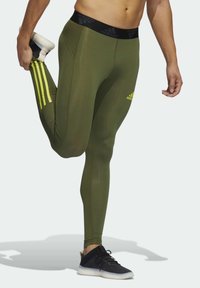 Olivegreenowe legginsy sportowe z czarnym pasem, z trzema pionowymi żółtymi paskami po bokach i dopasowanym krojem.