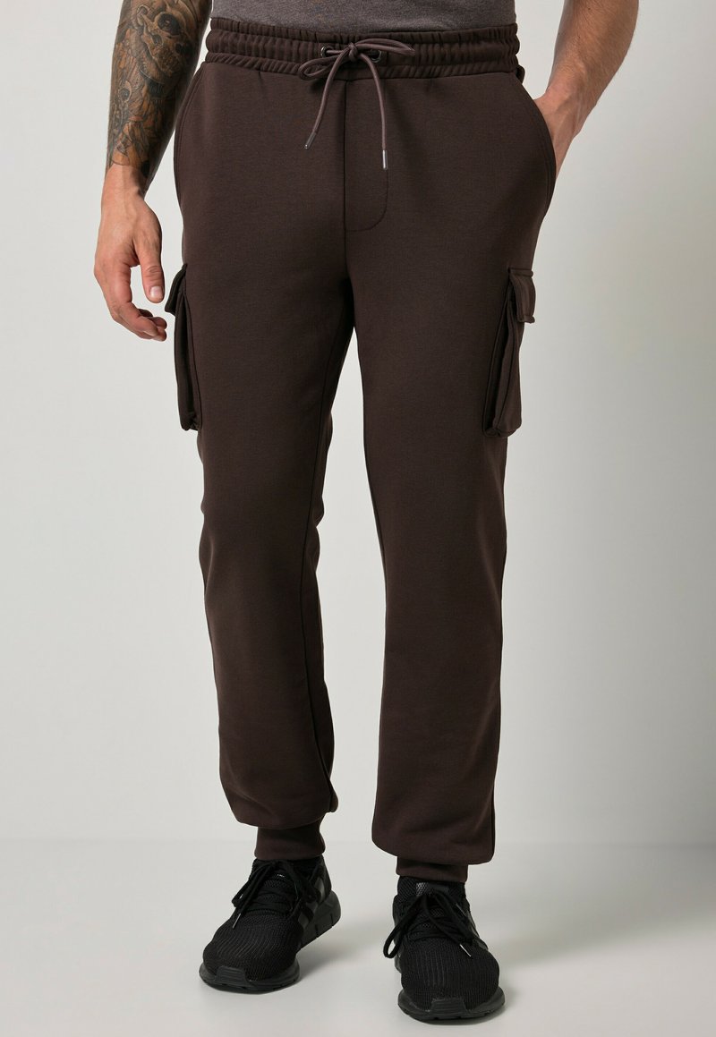 Homme portant un pantalon jogger cargo marron foncé avec taille à cordon et des baskets noires, debout devant un fond clair uni.