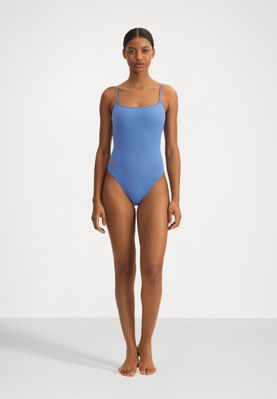 Maillot de bain une pièce bleu clair avec des bretelles fines, texture lisse, léger éclat et un petit logo rouge sur la hanche gauche.