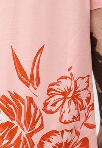 Hellrosa Stoff mit kräftigen roten tropischen Blatt- und Blumenmustern sowie dem Schriftzug "OAKLEY", getragen am Arm einer Person, die am Rand sichtbar ist.