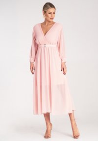 Maxi sukienka w delikatnym różu, z plisowanym dekoltem w kształcie litery V, długimi rękawami, przewężoną talią z metalowym paskiem oraz zwiewną, przezroczystą spódnicą.