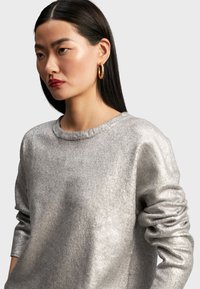 Metallisk silvertröja med lång ärm och rundad halsringning, med en texturerad yta och lätt rullade ärmslut.