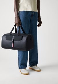 Black duffel bag with a smooth finish, featuring a front flap, a strap, red and white accent stripes, and sturdy dual handles.