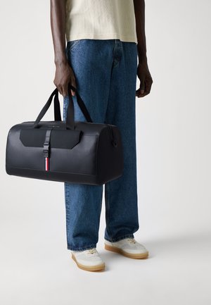 Sac de sport noir à la finition lisse, avec un rabat à l'avant, une bandoulière, des bandes d'accent rouge et blanc, et de solides doubles poignées.