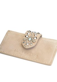 Tarjetero de ante beige que presenta tachuelas metálicas decorativas y piedras preciosas, con un logotipo en relieve. Textura suave con un detalle destacado.