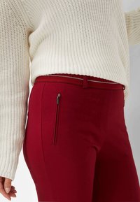 Red fitted pants with a smooth texture feature a side zip pocket and a contrasting waistband. Paired with a white knitted sweater.