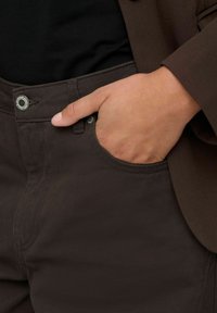 Pantalons en coton marron foncé avec une texture lisse, dotés d'une poche avant et d'une fermeture à bouton en métal. La taille est légèrement texturée.