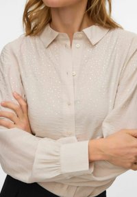 Helebeežikas heledast materjalist nööbitav särk kraega, millel on esiküljel helendavad aktsendid ning volüümikad varrukad kogutud mansettidega.