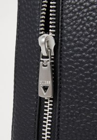 Svart texturerad läderdragkedja med en silverfärgad metalldragare försedd med "GUESS"-gravyr. Dragkedjans tänder har växelvis färg, vilket förstärker kontrasten.
