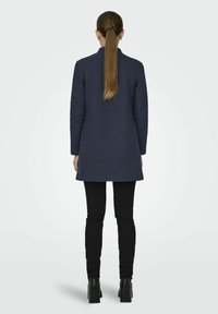 Cappotto blu navy con taglio dritto, maniche lunghe e colletto alto. Indossato sopra pantaloni neri e stivali neri Chunky. Tessuto testurizzato.