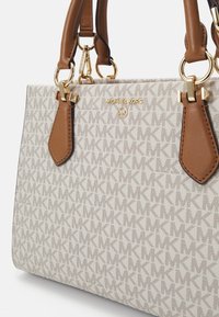 Beige canvas handtas met MK-monogram patroon, bruine leren handvatten, gouden accenten en logodetails. Heeft een gestructureerde vorm en accenten.