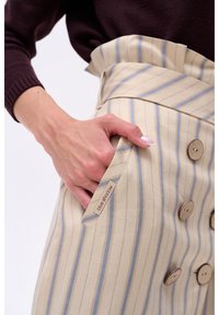 Beige gestreepte rok met blauwe en rode lijnen, voorzien van grote houten knopen en een zijzak gemarkeerd met een label "blue shadow."