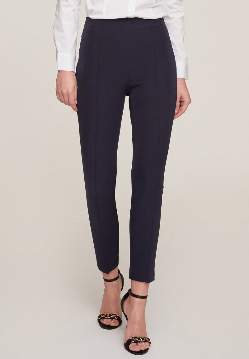 Femme portant un pantalon bleu marine sur mesure à longueur de cheville, avec une chemise blanche et des sandales à talons noirs ornées d'une chaîne dorée.