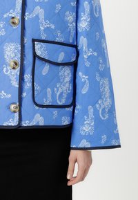 Quilted blue jacket featuring white floral and paisley patterns, with a front pocket trimmed in navy and round buttons.
