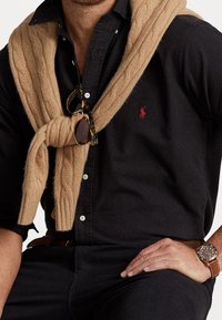 Svart skjorta med knappar och små prickar, kamelfärgad kabelstickad tröja över axlarna, bågar i sköldpaddsmönster solglasögon och en brun läderarmbandsklocka.