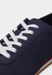 Σκούρο μπλε αθλητικό παπούτσι με λευκά κορδόνια, υφή στο πλάι, το λογότυπο "GUESS" ανάγλυφο στο πλάι και κόκκινη και λευκή ρίγα στη σόλα.