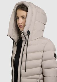 Puffer jacket in light beige with a thick, oversized hood, zip closure, drawstring accents, and quilted texture on the body.