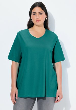 Donna con una maglietta verde acqua a scollo a V e maniche corte e pantaloni grigi, in piedi davanti a uno sfondo bianco semplice.