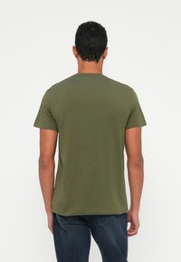 Vyras su trumpais garbanotais plaukais, vilkintis alyvų žalios spalvos trumpą rankovę marškinėlius ir tamsiai mėlynus džinsus, stovi prieš paprastą šviesų foną.