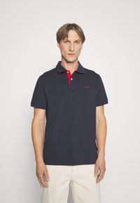Polo majica tamnoplave boje s kontrastnim crvenim ovratnikom, koristeći dvostruki gumb za kopčanje i izvezenim logotipom na prsima. Tkanina izgleda glatko i mekano.